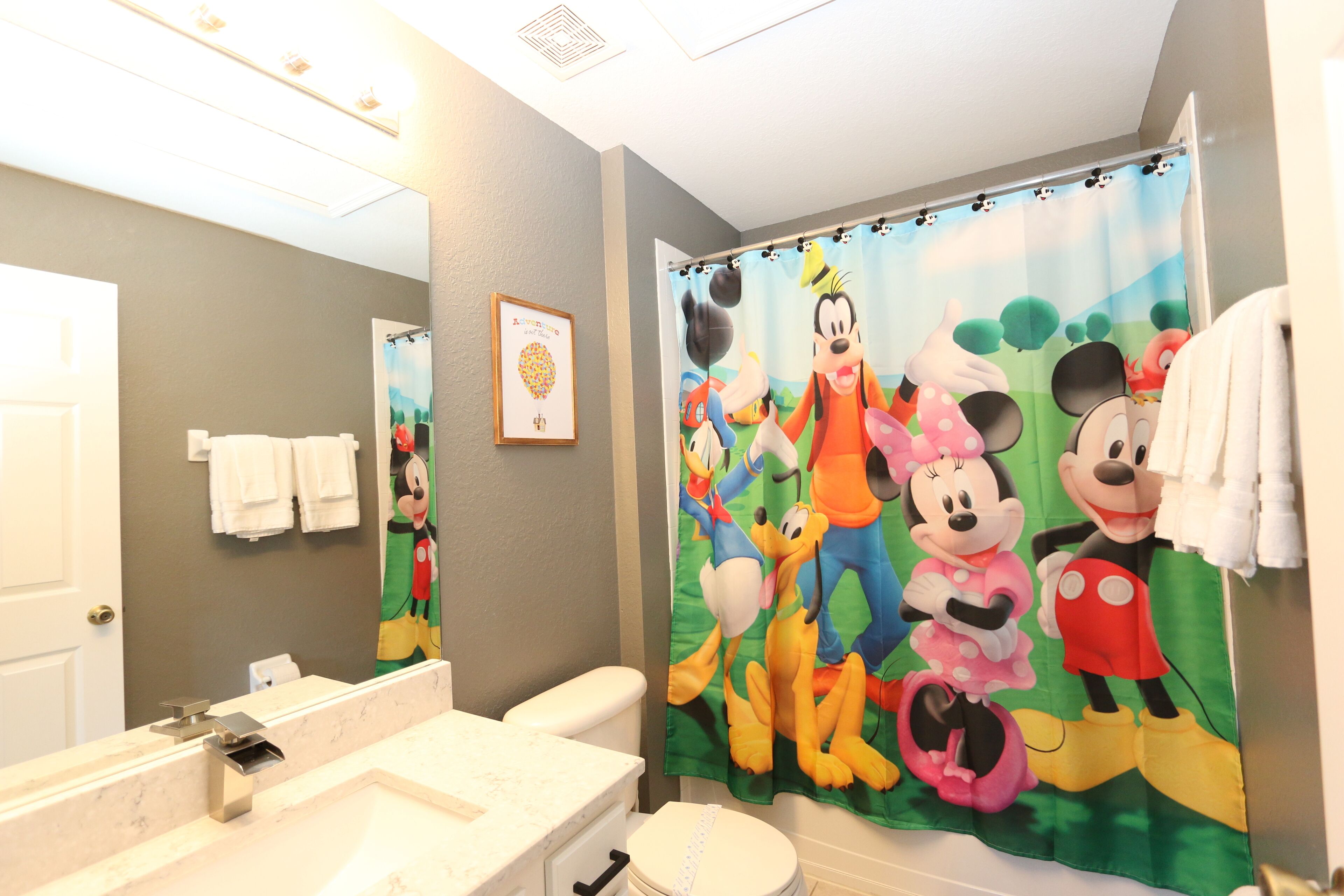 Un bany amb cortina de bany amb tema de Mickey Mouse, un vàter blanc i un lavabo de marbre.