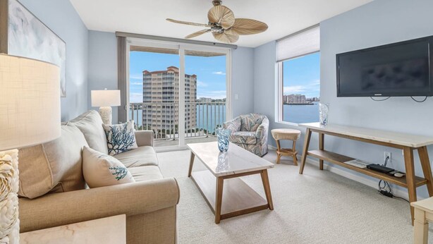 A modern living room with a sofa, coffee table, and armchair, a TV, and a balcony with a city view. - Beautiful 1-bedroom condo with the best views ocean,back bay & beautiful sunset (Fort Myers Beach)