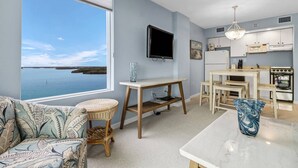 A modern living room with a large window overlooking a body of water, a television, a dining area with a table and chairs, and a kitchen with white cabinets and appliances. - Beautiful 1-bedroom condo with the best views ocean,back bay & beautiful sunset (Fort Myers Beach)