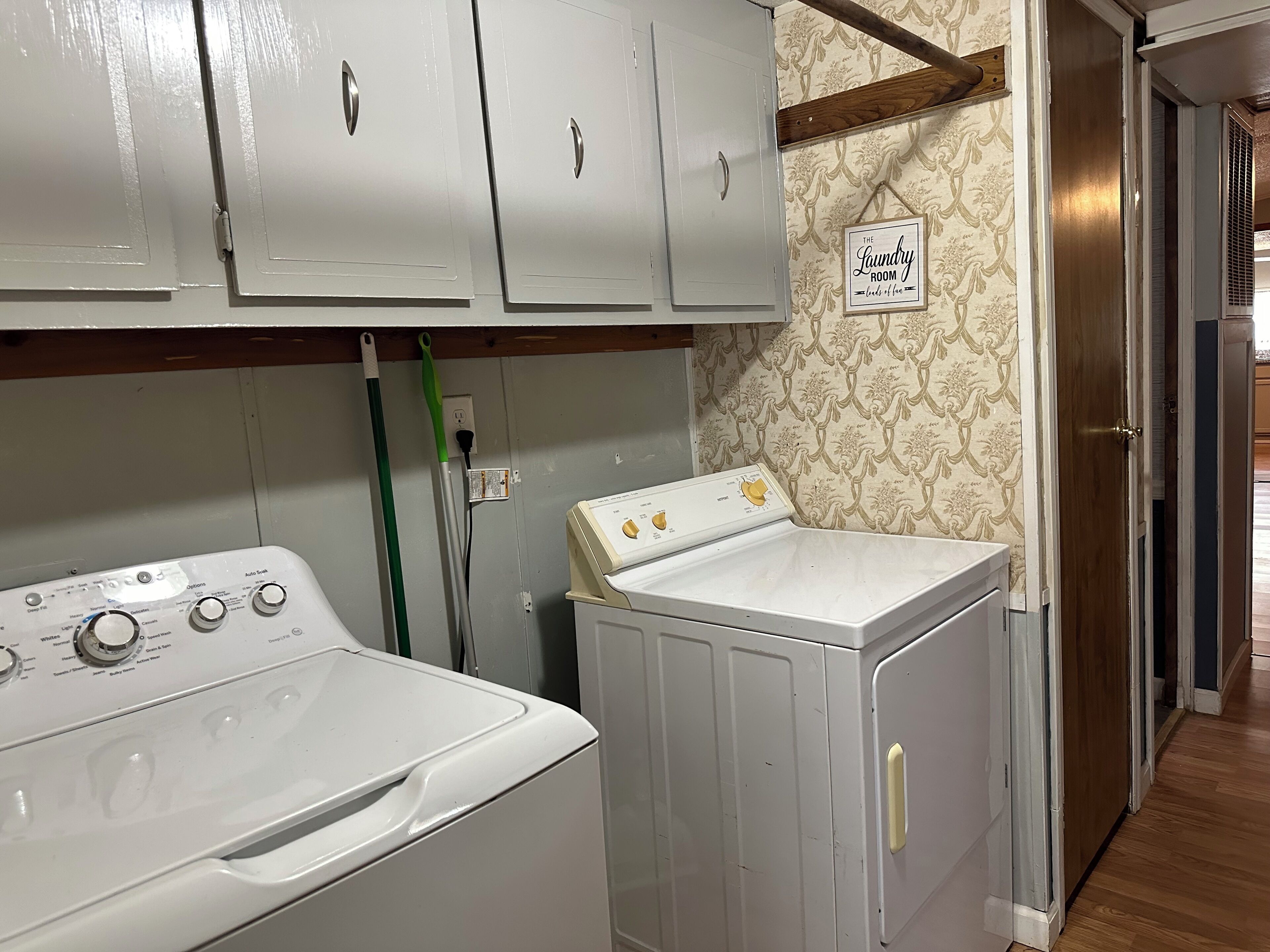 A laundry room with a washing machine and dryer, wooden cabinets, a green mop, and a sign that reads "The Laundry Room."