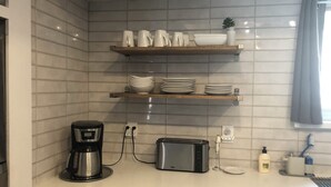 Eine Küche mit weiß gefliesten Wänden, einer Kaffeemaschine, einem Toaster und einem Regal mit weißen Geschirr und Schüsseln.