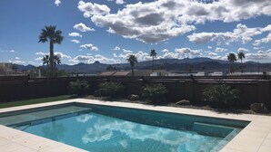 Ein rechteckiger Swimmingpool mit klarem, blauem Wasser, umgeben von einer gefliesten Liegefläche. Im Hintergrund sind Palmen und ein Gebirge unter einem bewölkten Himmel zu sehen.