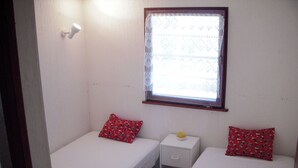 A small, single-room accommodation with two single beds, each with a red patterned pillow. There is a small bedside table with a yellow object on it, and a window with a lace curtain.