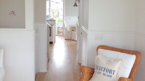 A spacious hallway with a wicker rocking chair and a white cushion with text, leading to a kitchen area.