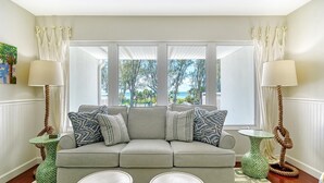 Ein Wohnzimmer mit grauem Sofa, gemusterten Kissen, einem grün-weißen Beistelltisch, einem weißen Couchtisch und Blick ins Freie durch große Fenster.