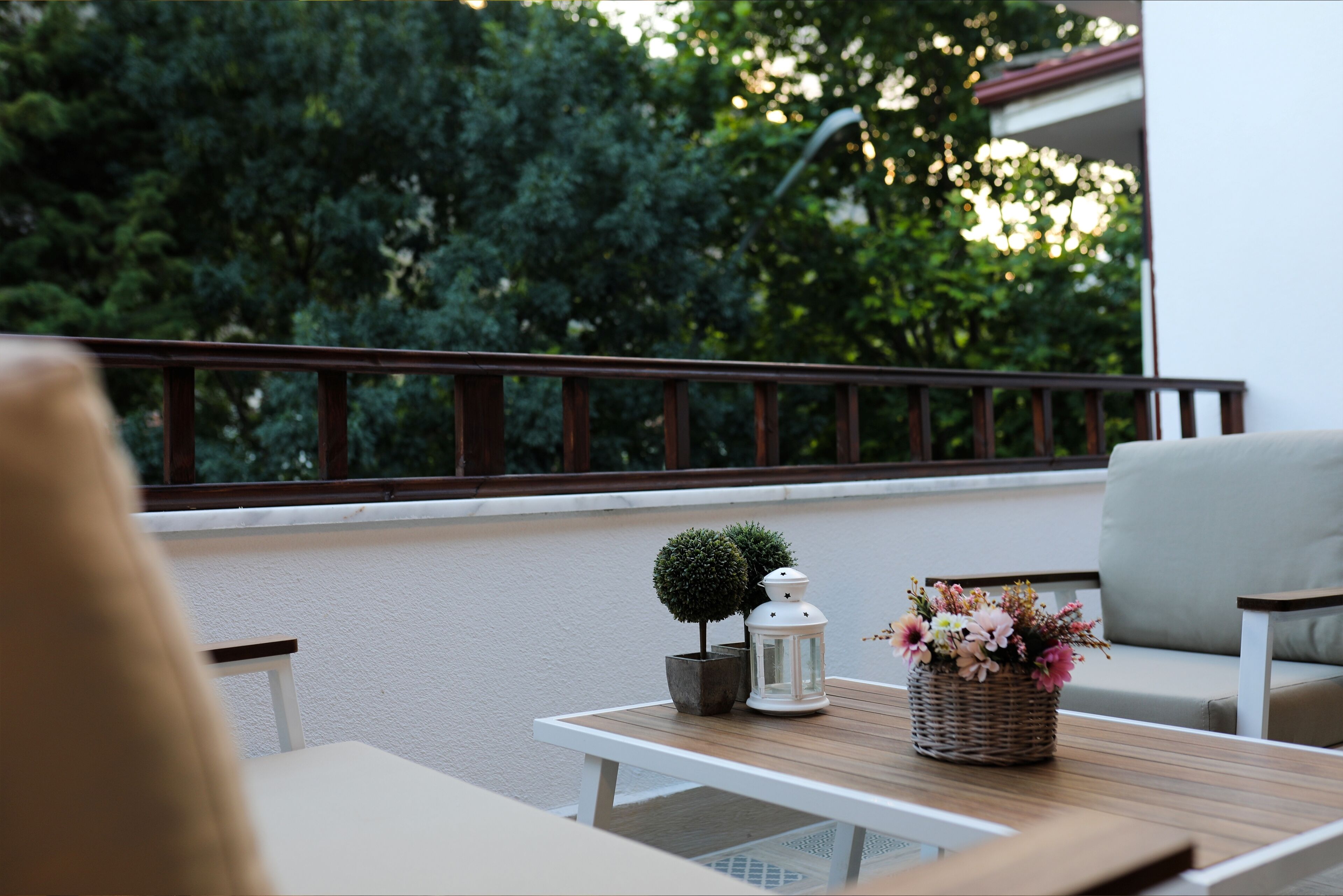 A balcony with a table, chairs, a small potted plant, a lantern, and a basket of flowers.