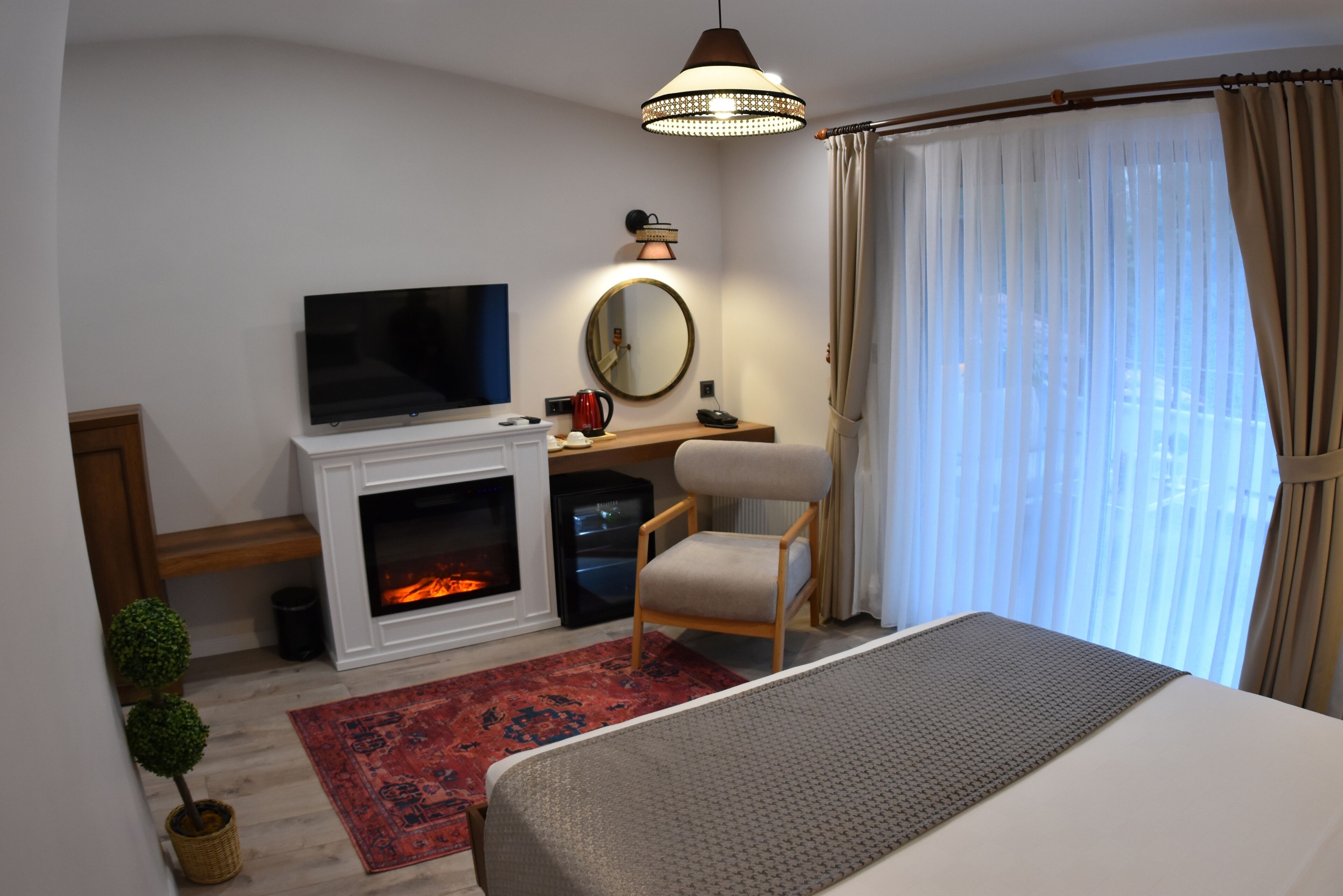 A hotel room with a television, fireplace, desk, chair, and large window with curtains.