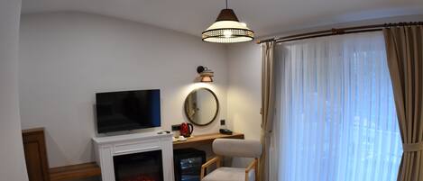 A hotel room with a television, fireplace, desk, chair, and large window with curtains.