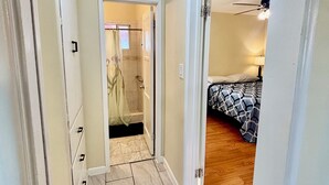 Un couloir mène à une salle de bain avec douche et à une chambre à coucher équipée d’un lit et d’un ventilateur de plafond.