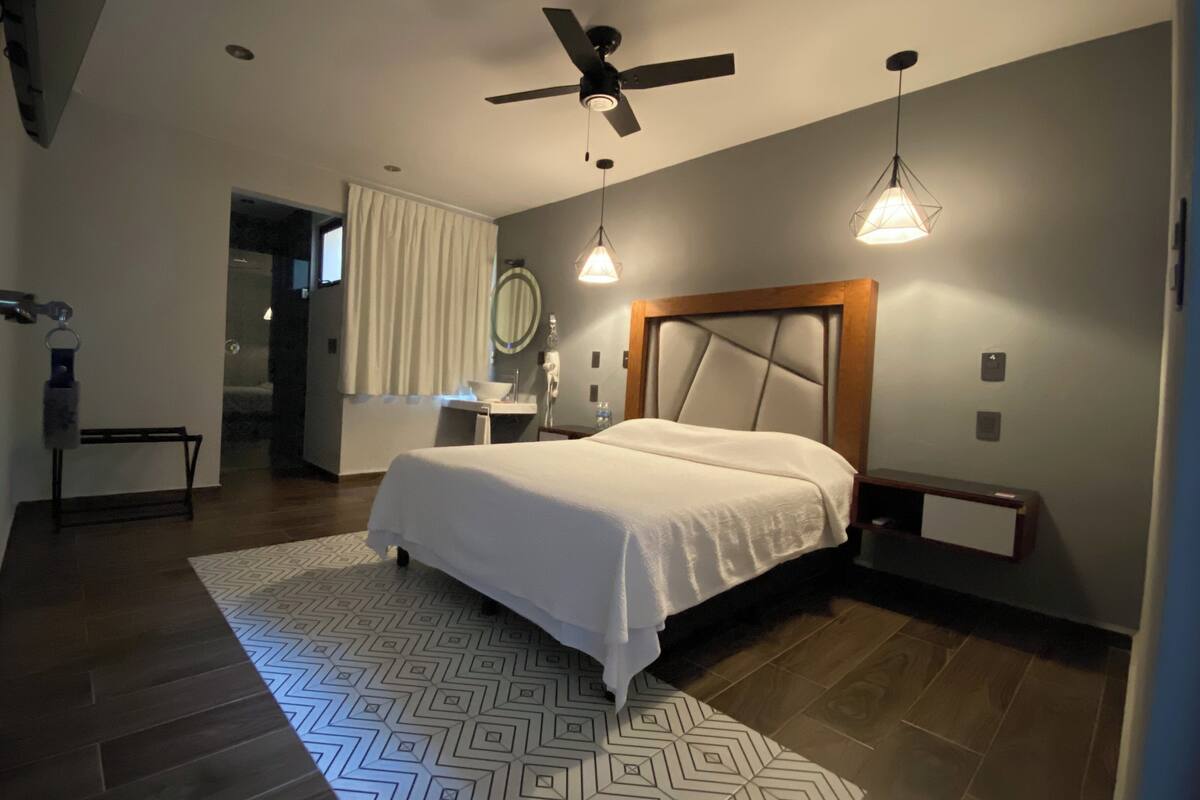 A modern hotel room with a large bed, a ceiling fan, a bedside table, a nightstand, a mirror, a bathroom with a sink and a toilet, and a patterned rug.