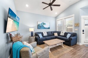 A living room with a sectional sofa, a coffee table, a TV mounted on the wall, and a ceiling fan.