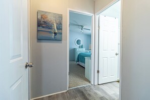 Um corredor leva a um quarto com ventilador de teto, uma cama com colcha azul e um quadro de um veleiro na parede.