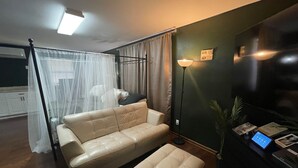 Un salon avec un canapé et un pouf de couleur beige, une lampe sur pied, un téléviseur et une petite table sur laquelle sont posés des livres et un appareil.