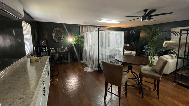 Une pièce comprenant une table à manger, des chaises, un ventilateur de plafond, un lit avec des rideaux blancs et une grande plante.