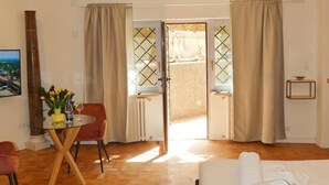 Une chambre d’hôtel comprenant un lit, une table à manger avec des chaises, une petite table avec un vase et un verre à vin, et une vue sur l’extérieur par une porte.