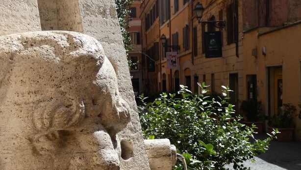 Une fontaine en pierre dont l’eau jaillit de sa gueule, entourée d’une rue pavée et de bâtiments.