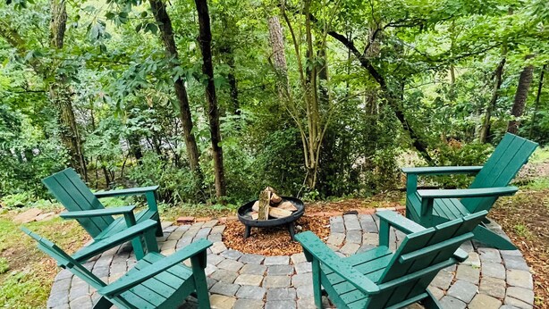 Een ronde vuurkuil van steen, omringd door vier groene Adirondack stoelen in een bebost gebied.