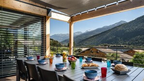 A dining table set on a balcony with a mountain view, surrounded by chairs and various dishes and drinks. - CHALET DES EPICURIENS (AIME LA PLAGNE)