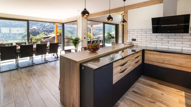 A modern kitchen with a large island, wooden flooring, and a view of a mountainous landscape through large windows. - CHALET DES EPICURIENS (AIME LA PLAGNE)