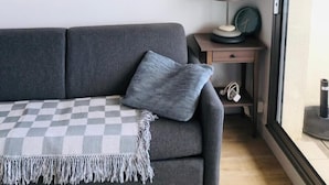 Una sala moderna con un sofá gris, una manta de cuadros, un cuadro enmarco de un paisaje marino, una mesita de noche con una lámpara y vistas a través de una puerta de cristal.
