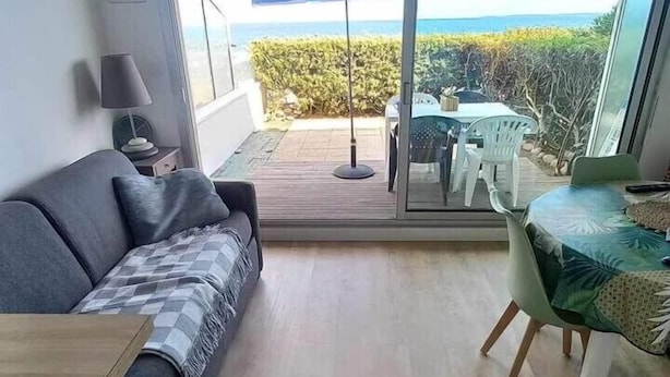 Una sala de estar moderna con un sofá, una mesita redonda y un comedor con mesa y sillas. Hay un ventanal con vistas al océano y un área de patio con sombrilla.