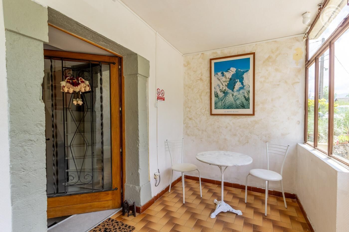 Una habitación pequeña con una puerta de madera, una puerta de cristal, una mesa redonda con dos sillas, un cuadro en la pared y una ventana.