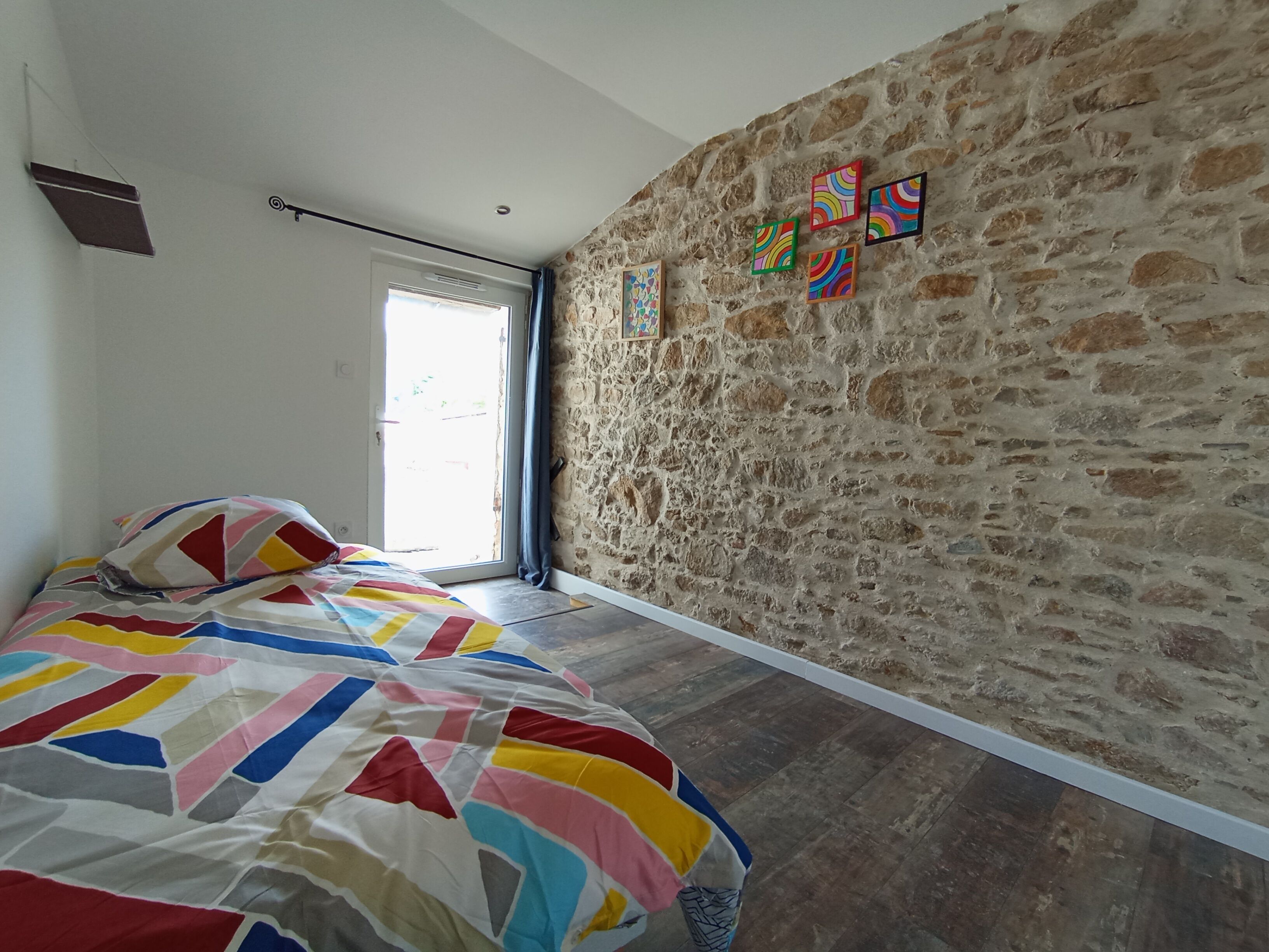A bedroom with a stone wall, a bed with a colorful quilt, and a window with curtains.
