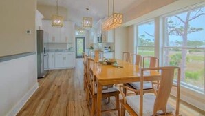 Un comedor amplio con una larga mesa de madera y sillas, una cocina al fondo y ventanas grandes que ofrecen vistas al exterior.