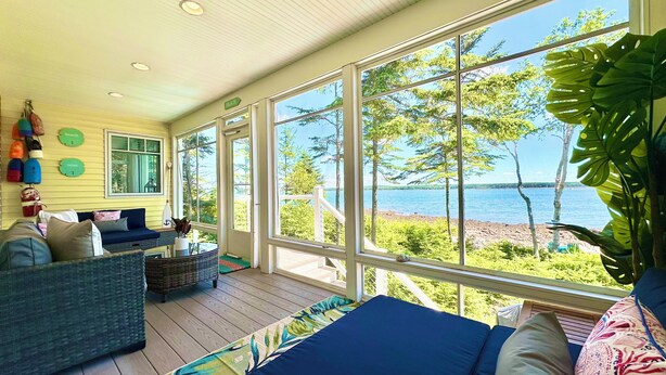 A sunlit room with a view of a lake, furnished with a sofa, a chair, and a small table. - ⭐NEW⭐ Stunning Beachfront Living w/ Gameroom & Firepit Near Acadia  (Gouldsboro)