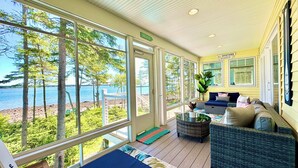 A sunlit room with large windows overlooking a beach, furnished with a sofa and a dining table. - ⭐NEW⭐ Stunning Beachfront Living w/ Gameroom & Firepit Near Acadia  (Gouldsboro)
