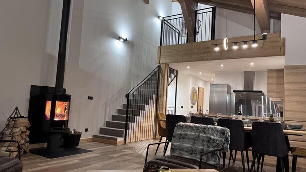 Un amplio salón con techo alto, chimenea, mesa de comedor preparada para cuatro comensales y una escalera que sube a un nivel superior.