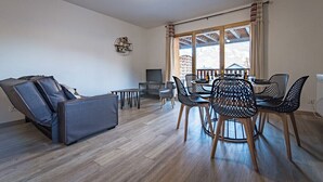 Ein modernes Wohnzimmer mit Massage-Sessel, Esstisch mit Stühlen und großem Fenster mit Vorhängen.