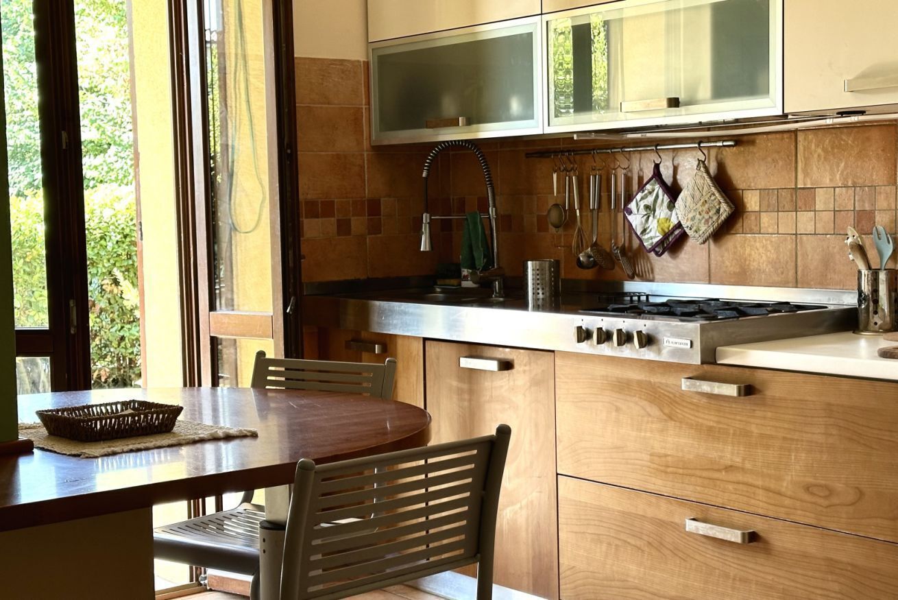 A kitchen with a stove, sink, and wooden cabinets. There is a window with a view of greenery outside.