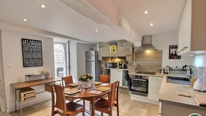 A modern kitchen with a central island, built-in oven, and a stone wall. There is a dining area with wooden chairs and a table, and a large window providing natural light. - La Maison Gousset - GUINGAMP (GUINGAMP)