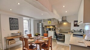 Une cuisine moderne dotée d’un îlot central, d’un four encastré et d’un mur en pierre. On y trouve un coin repas avec des chaises et une table en bois, ainsi qu’une grande fenêtre qui laisse entrer la lumière naturelle.