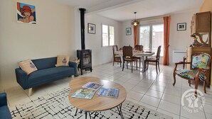 A living room with a sofa, a round coffee table with magazines, a dining table with chairs, a fireplace, and a large window with curtains. - Les 2 Pointes - PLOUEZEC (PLOUEZEC)