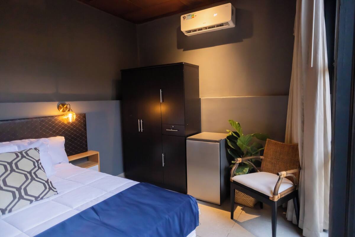 A bedroom with a bed, wardrobe, bedside table, chair, potted plant, and an air conditioning unit.