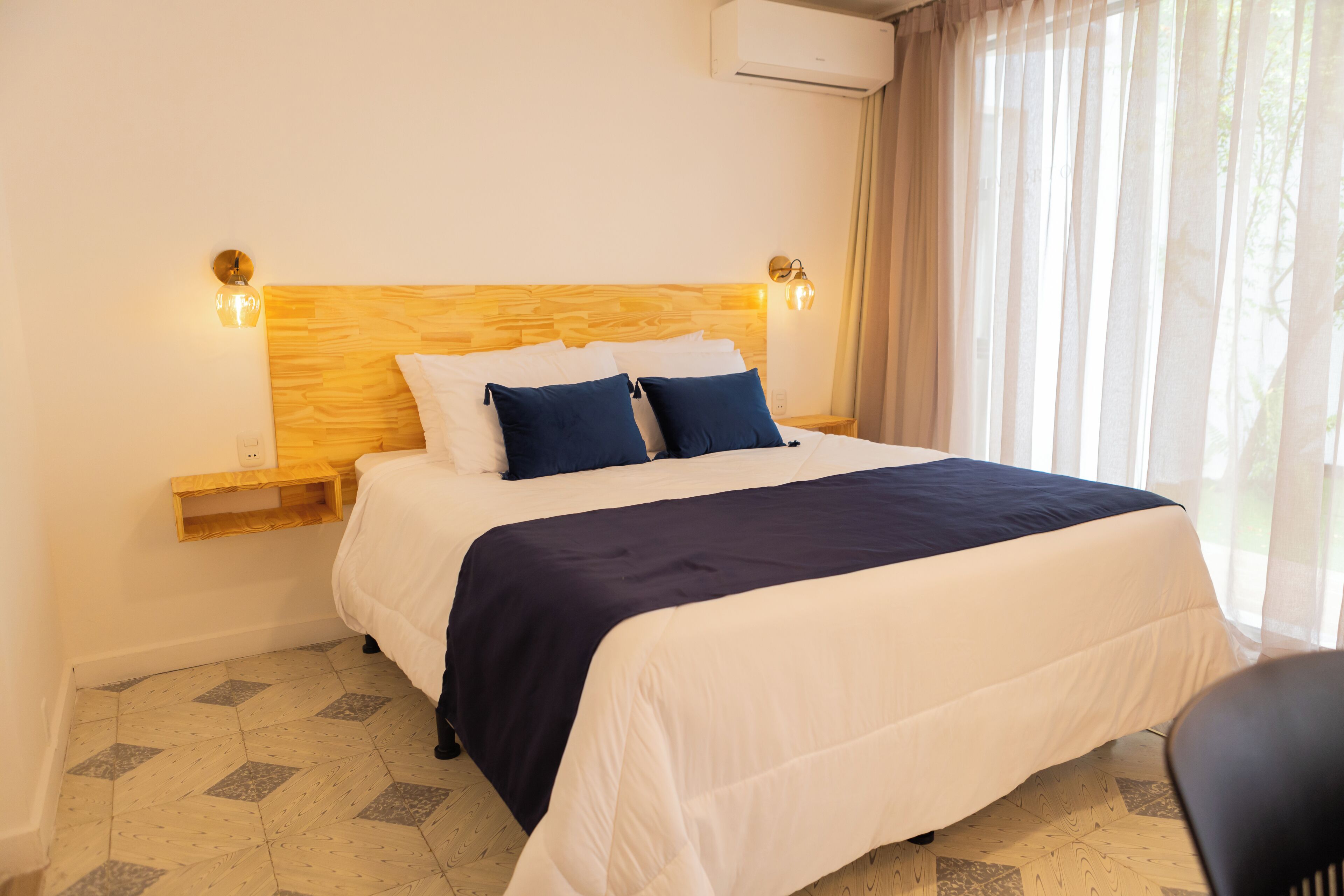 Una cama bien hecha con cabecera de madera, dos lámparas de noche y una ventana con cortinas traslúcidas.