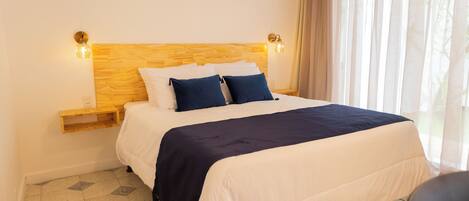 Una cama bien hecha con cabecera de madera, dos lámparas de noche y una ventana con cortinas traslúcidas.