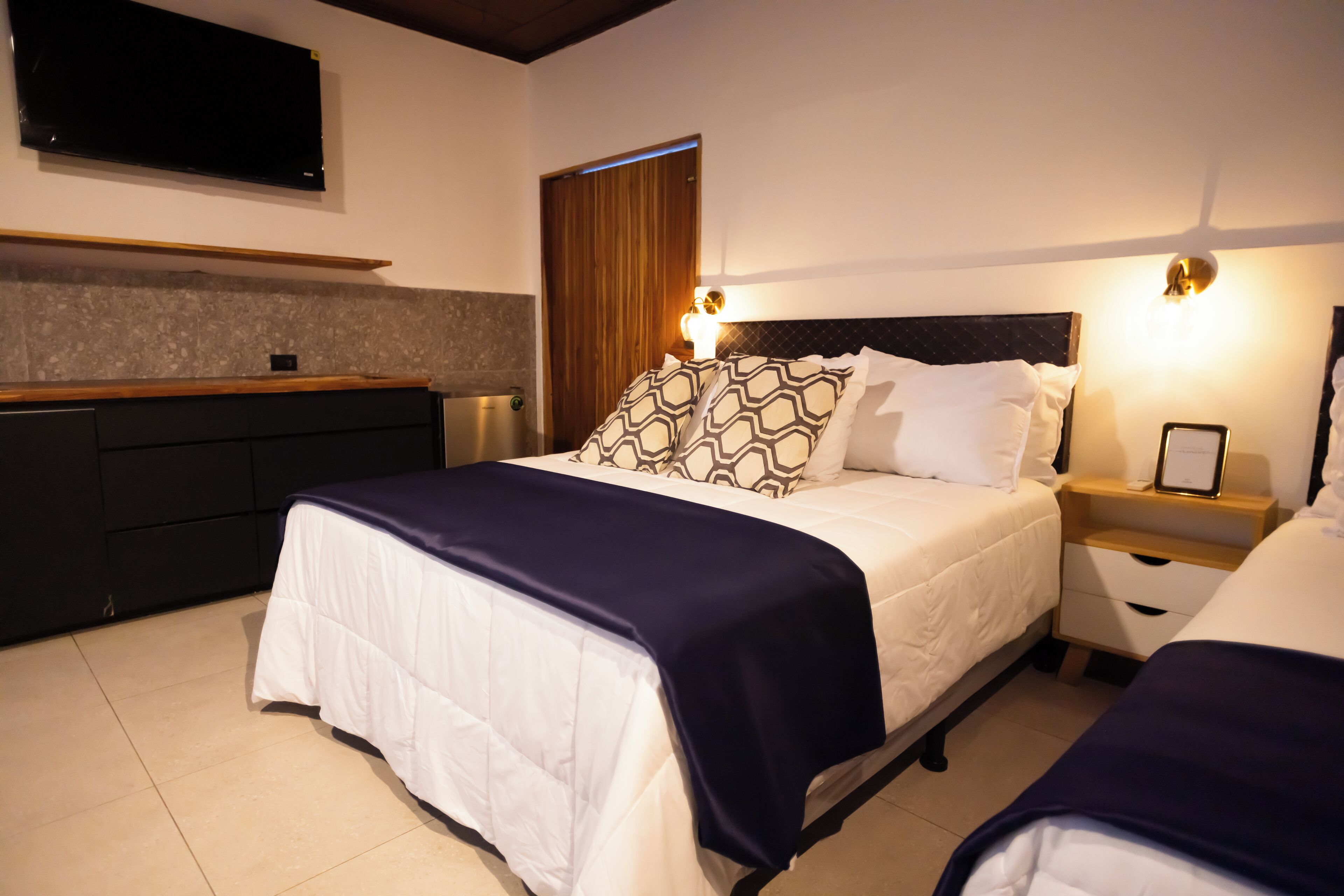 Une chambre d’hôtel avec un lit, des tables de chevet, une télévision, une étagère en bois et une fenêtre avec des rideaux.