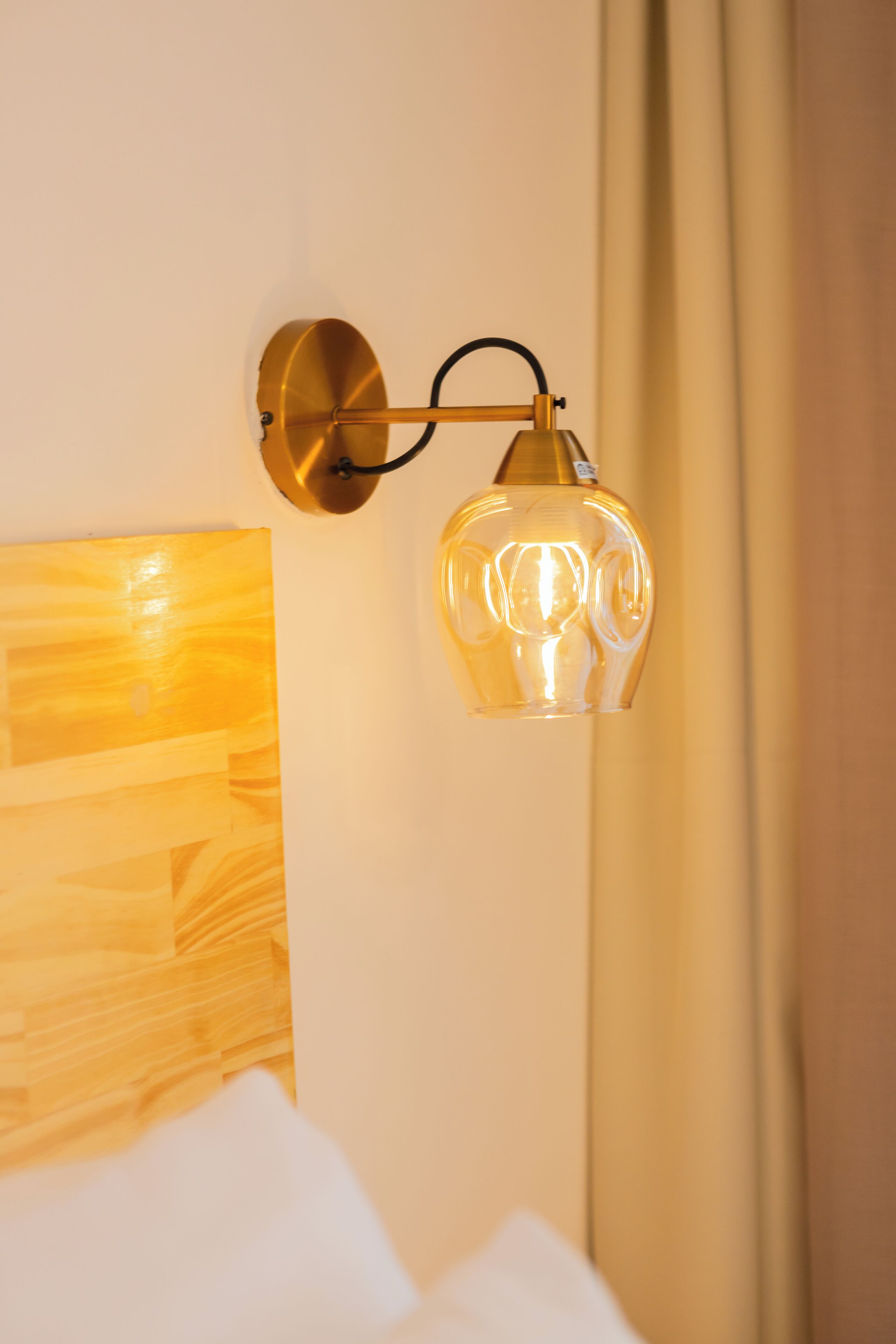 A wall-mounted lamp with a clear glass shade, a wooden headboard, and a curtain.