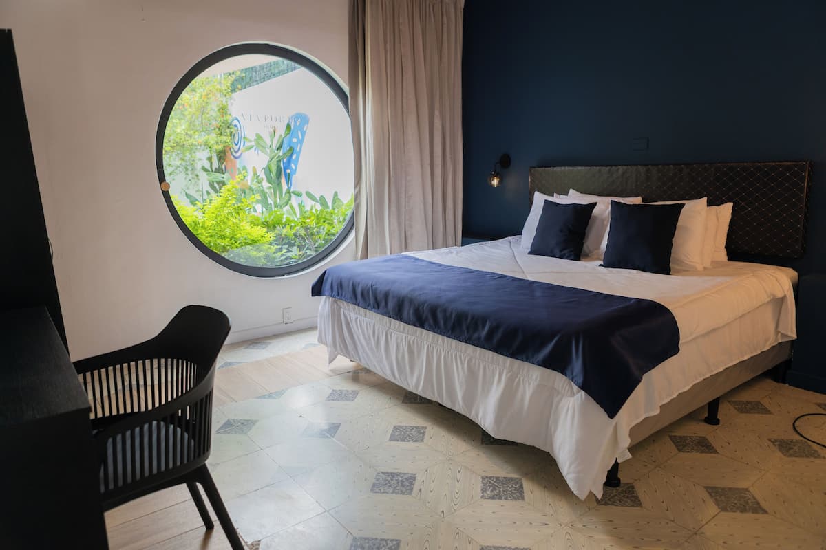 A bedroom with a bed, a round window, a chair, and a view of greenery.