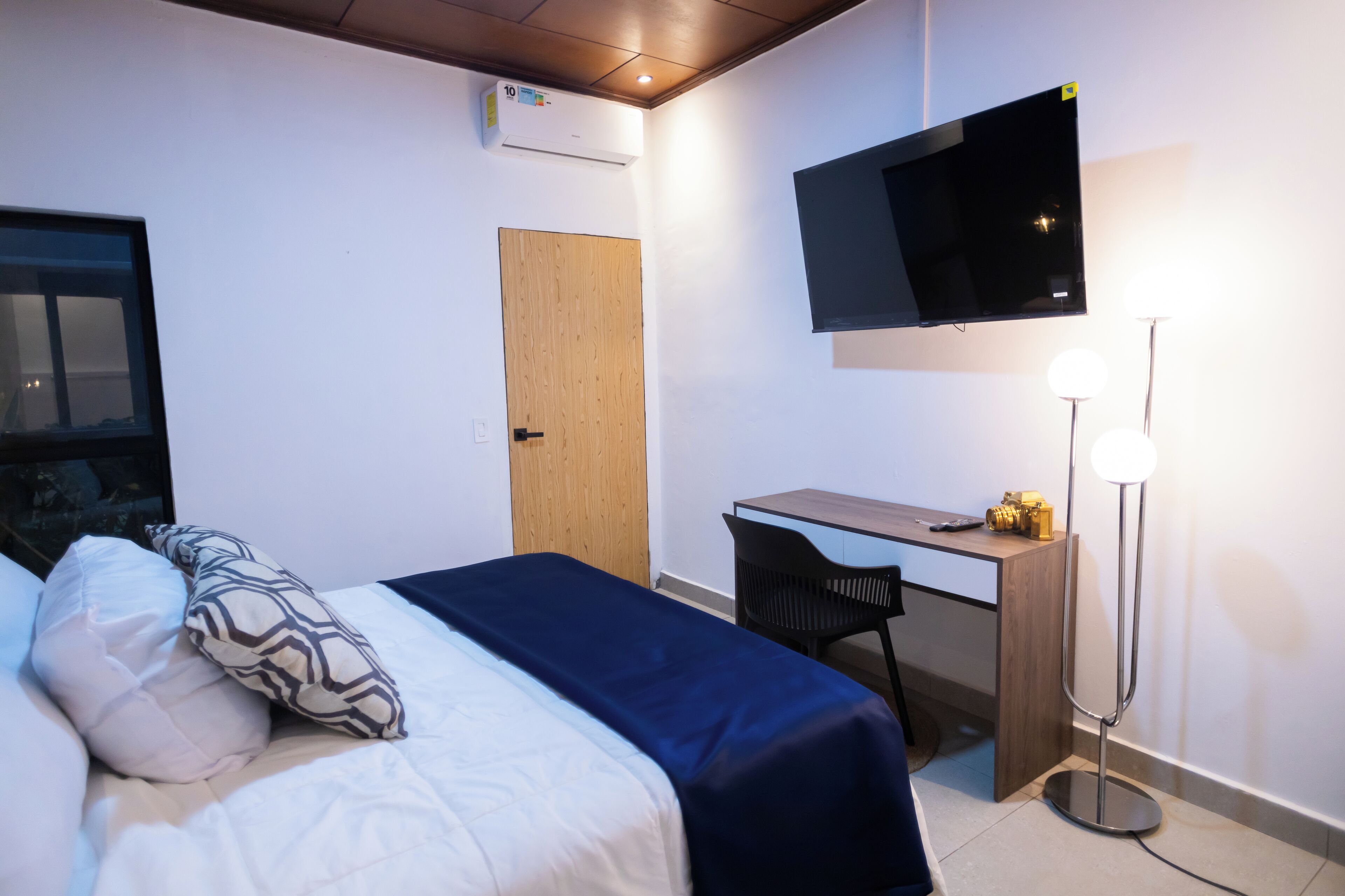A hotel room with a bed, a desk, a chair, a TV, and a standing lamp.