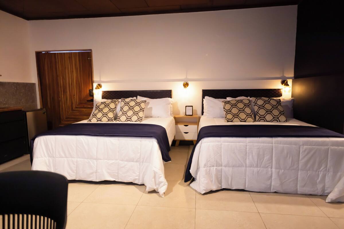 A hotel room with two beds, each with patterned pillows and a dark blue bedspread. There are bedside tables with lamps and a wooden door in the background.
