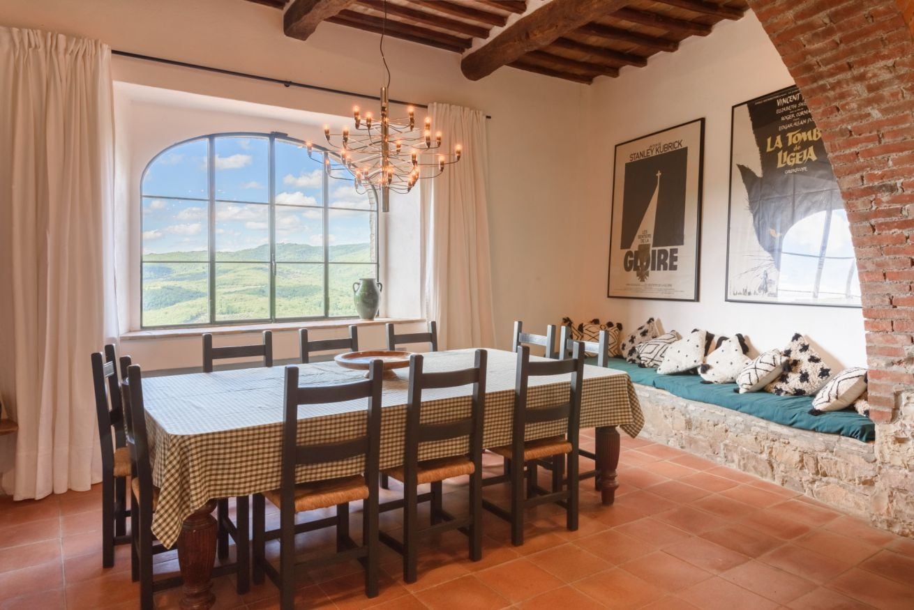 A dining area with a table set for six, a chandelier, a view through a large window, and a brick wall with a bench.