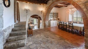 Un interior rústico con paredes de piedra, una escalera de madera, una zona de comedor con mesa y sillas, y una cocina visible a través de un arco.