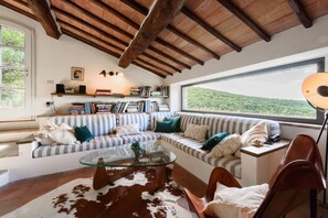 A cozy living room with a wooden ceiling, a glass coffee table, a striped sofa, and a view of a forest.