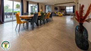 Un comedor amplio con una mesa larga y numerosas sillas, una sala de estar con sofá y sillones, y un ventanal con vistas al exterior.