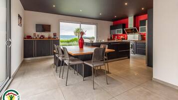 A modern kitchen with a central island, dining area with chairs, and a large window offering a view of the sea.