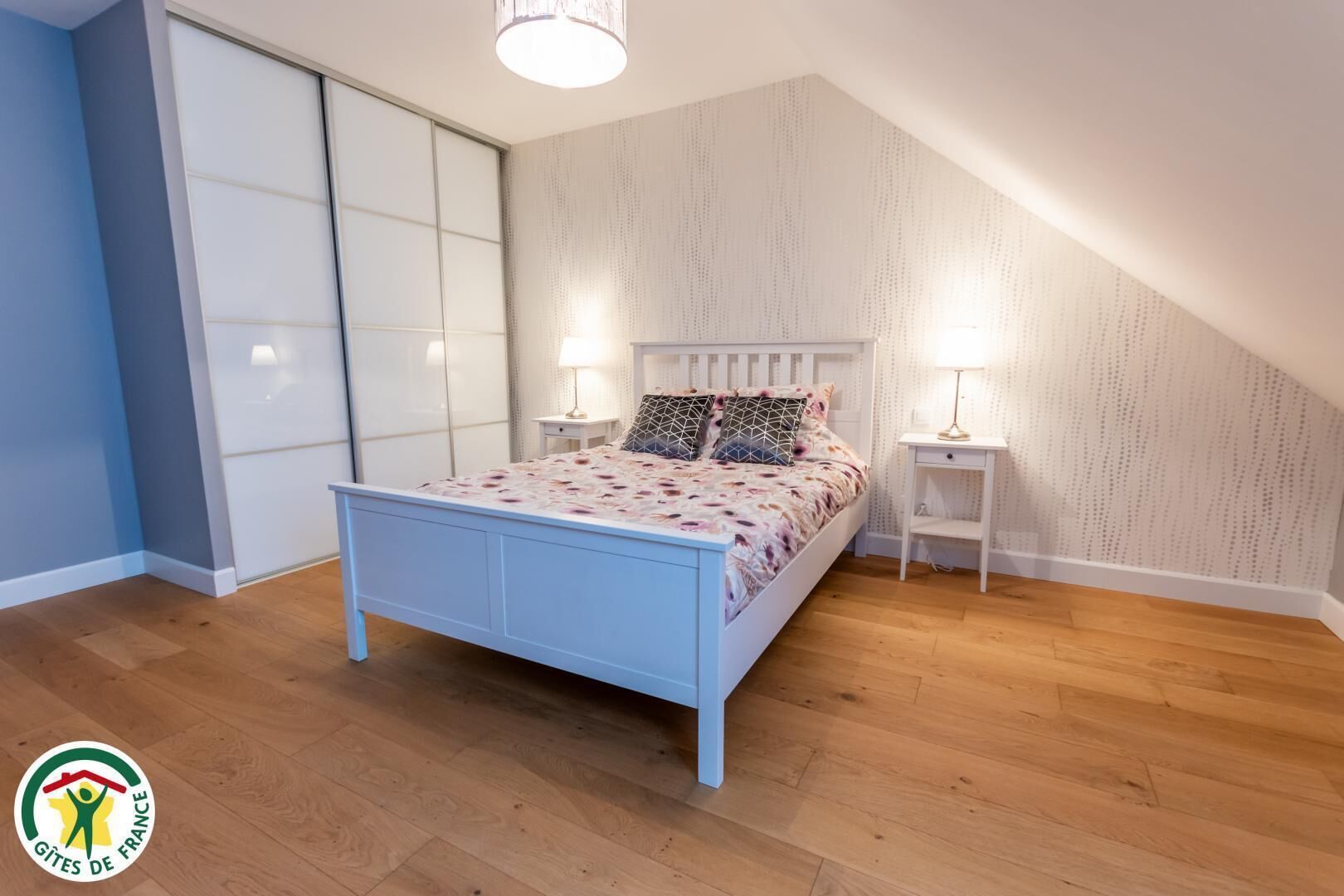 Una camera da letto con un letto bianco, due comodini con lampade, un armadio e un soffitto spiovente.